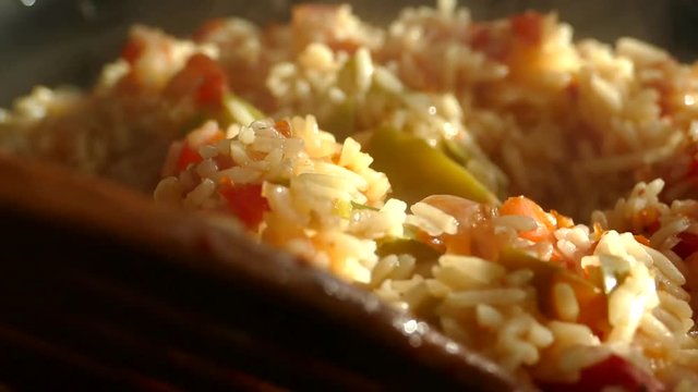 Prepare pilaf in a large frying pan. A dish of rice and pieces of vegetables. Steam comes from a hot lunch. Home kitchen. Pilaf is stirred with a wooden spatula.