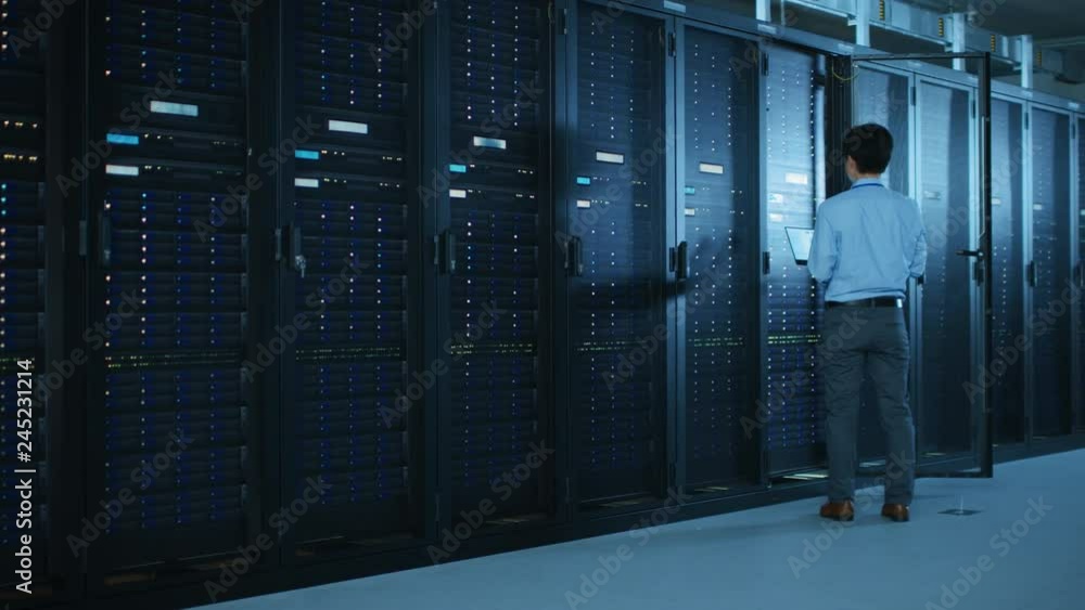 Poster In the Modern Data Center: IT Engineer Working with Server Racks, Uses Laptop for Maintenance and Diagnostics. Checking Stability of the System
