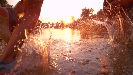 SLOW MOTION TIME WARP, SUN FLARE, CLOSE UP: Fit training partners run in refreshing river water towards beautiful summer sunset. Glassy drops of water fly as young couple jogs through serene nature.