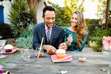 Smiling young multiethnic couple eating cupcake while sitting at table in backyard during cocktail party