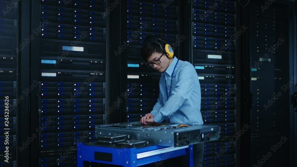 Poster In the Modern Data Center: IT Technician Wearing Protective Headphones Working with Server Racks, on a Pushcart Installing New Hardware. Engineer Doing Maintenance and Diagnostics of the Database.
