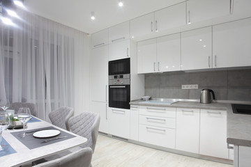 White kitchen interior with grey chairs and served table with white plates glasses and moss on the wall in new luxury home with lights on