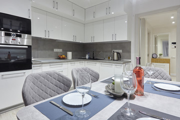 White kitchen interior with grey chairs and served table with white plates glasses and moss on the wall in new luxury home with lights on