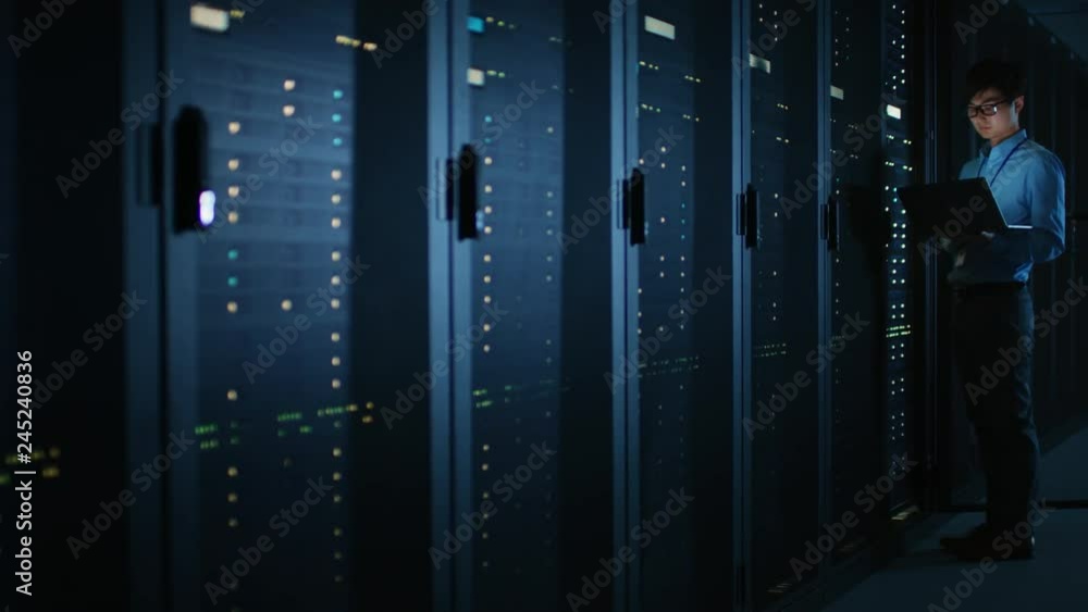 Poster In Dark Data Center: Male IT Specialist Stands Beside the Row of Operational Server Racks, Uses Laptop for Maintenance. Cloud Computing Artificial Intelligence Supercomputer Cybersecurity. Neon Lights