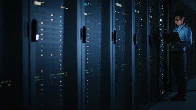 In Dark Data Center: Male IT Specialist Stands Beside the Row of Operational Server Racks, Uses Laptop for Maintenance. Cloud Computing Artificial Intelligence Supercomputer Cybersecurity. Neon Lights