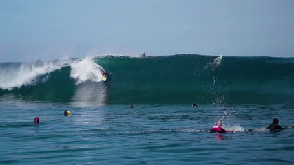 Slow motion male extreme pro surfer surfing a huge tube barrel wave at the tropical world famous surf location Teahupoo with crowds of people watching on a sunny day with blue crystal clear water