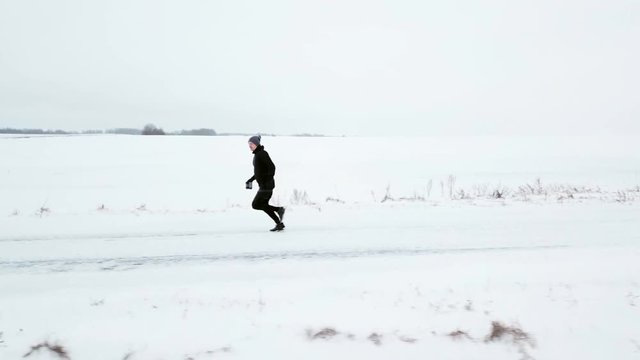 A man in black tracksuit is running along a snowy road. Winter, around the snow. White desert. Sport. Aerial view. Drone flies nearby with a man