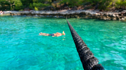 Naklejka premium The tourist snorkeling at stone coast of Brock island at sea in Croatia. Boat tour entertainment. Selective focus on the anchor rope of the arrived boat. Nautical and marine vacation background.