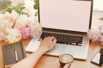Girl hands on stylish laptop with empty screen and coffee, phone, black notebook and peonies on...