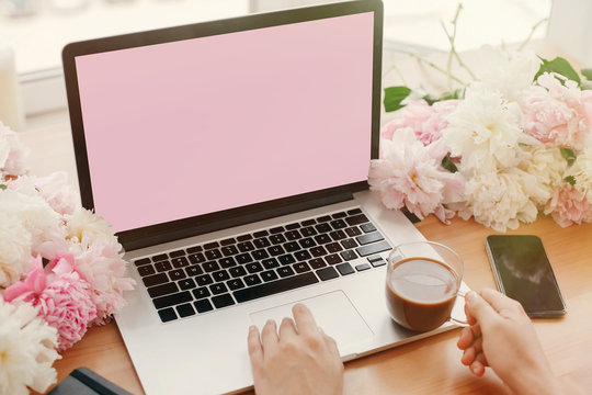 Girl hands on stylish laptop with empty screen and coffee, phone, black notebook and peonies on rustic wooden table. Freelance concept. Workplace. Space for text. Women's Day. - Powered by Adobe