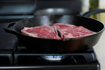 New York strip steaks cooking in a cast iron pan on a natural gas stove top.
