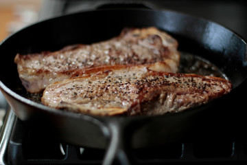 New York strip steaks frying in a cast iron pan on a natural gas stove top.