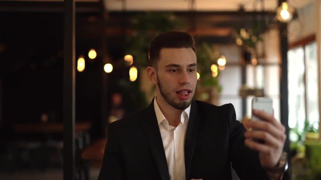 Young charming man is sitting in a cafe indoors and talking in a video chat. Man is holding a phone in front of his face, looking directly at phone's camera and smiling.
