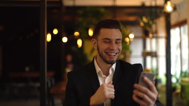 Young man is sitting in a cafe indoors, talking with somebody in a video chat, showing him his braces and pointing on them. Man is smiling and laughing out loud.