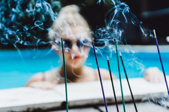 Blurred background of aromatic sticks and smoke with young beautiful blonde woman in sunglasses with a good figure with red lips make-up posing in a pool of blue water. Outdoor portrait close up. 