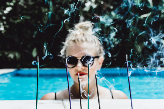 Blurred background of aromatic sticks and smoke with young beautiful blonde woman in sunglasses with a good figure with red lips make-up posing in a pool of blue water. Outdoor portrait close up. 