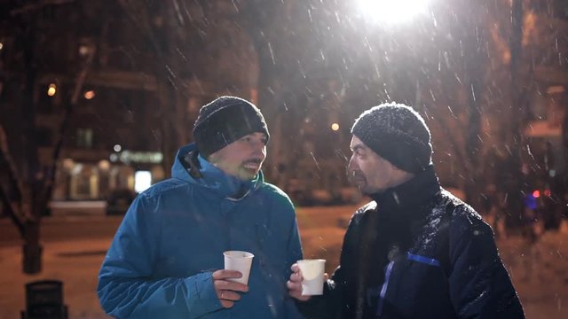 Two young men stand on the street in winter, heavy snow, chatting and drinking coffee