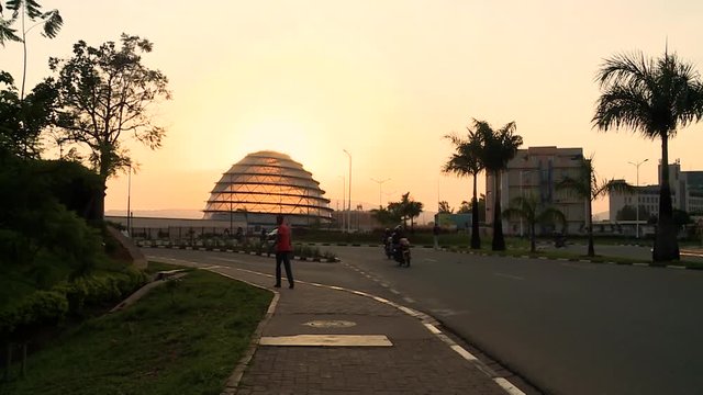 Very wide view of a Kigali roundabout at sunset, cars and people, convention center dome in front of the sun, Rwanda.