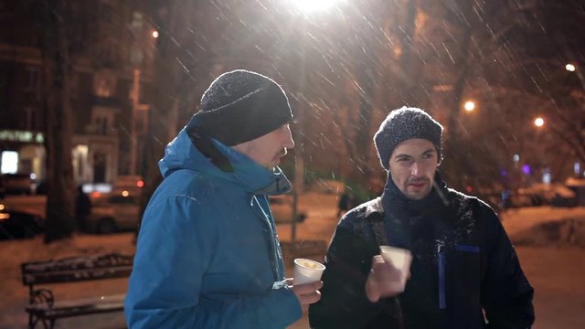 Two young men stand on the street in winter, heavy snow, chatting and drinking coffee