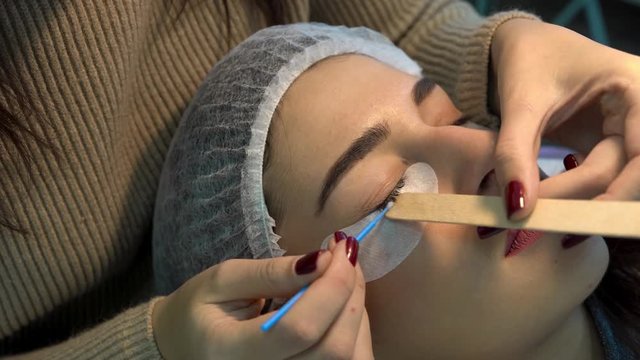 Eyelahes extension concept. Female expert smoothing and extenting female client eyelashes using special cotton stick and wooden stick. The female client lying on couch with eyes closed in salon/spa