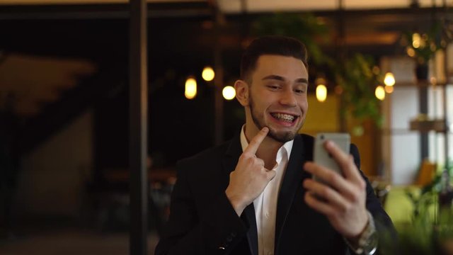Young good-looking man with a beard is wearing a business style suit, sitting in a cafe indoors, talking with somebody in a video chat, showing him his braces and pointing on them.