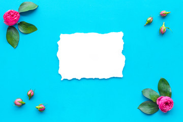 Floral mockup. Flowers, petals, leaves around white paper on blue background top view copy space frame