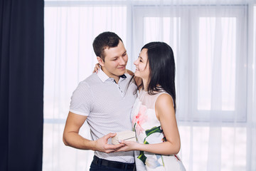 Naklejka premium Beautiful young couple in love man and woman man gives a gift in a box on the background of the window