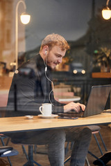 one young happy man, sitting in coffee shop and using his laptop, typing on keyboard, looking to computer screen. Shoot thought window outside (with reflections).