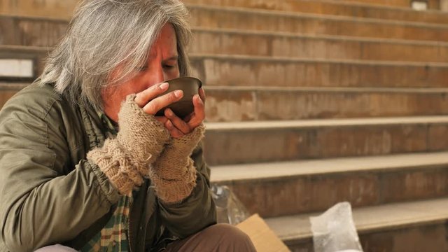 Homeless old man eating soup received from volunteers while sitting on stairs in the city