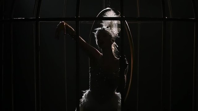 Graceful girl gymnast in bird costume riding a hoop in a cage . Black background. Slow motion. Close up. Silhouette