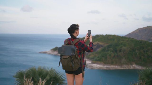 Woman traveler with backpack admiring sea view and making photo on the shore of a tropical beach sunset using smartphone. Female wearing red checkered shirt and sunglasses.