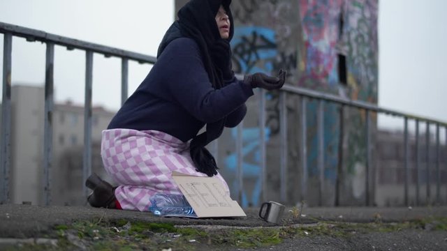 Adult homeless woman with outstretched hand sits on the bridge in cold windy grey weather asking for alms and help and one woman stopped to put coins