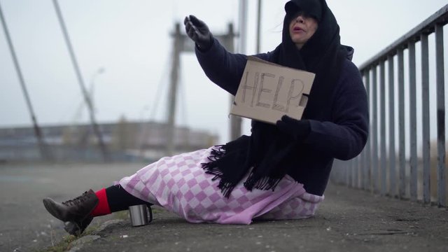Adult homeless woman sits on the bridge in cold windy grey weather asking for alms and help