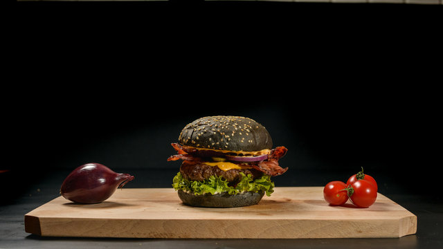 An appetizing black hamburger on a wooden board with fresh vegetables.