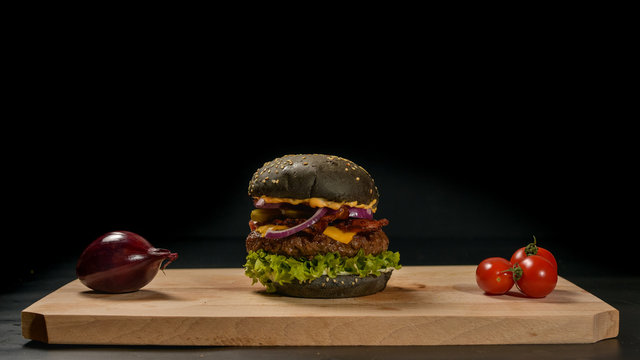 An appetizing black hamburger on a wooden board with fresh vegetables.