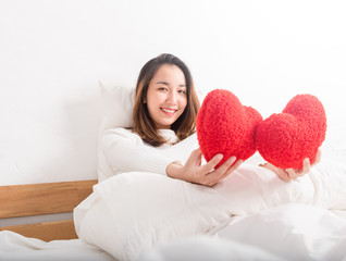 Obraz na płótnie Canvas Beautiful Asian Happy woman holding two red heart-shaped pillow on a bed in a white bedroom.Falling in love moment,valentines day concept