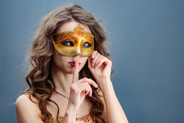 Woman in a golden dress and carnival mask on a blue background touching her finger to her lips