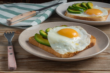 Toasts bread with fried eggs, avocado and cucumber slices on plates and wooden table with fork and knife. Soft focus.