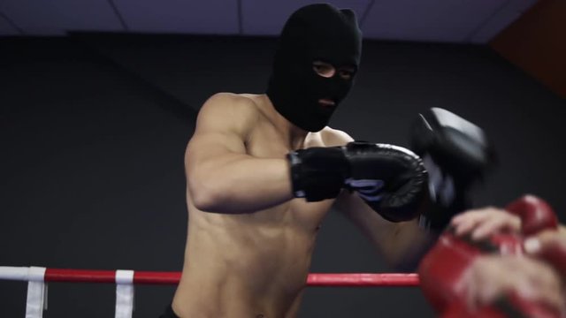Aggressive boxer works with boxing paws with his coach in the ring. Practicing a series of leg punches. Shirtless athlete in black boxing gloves and thief mask