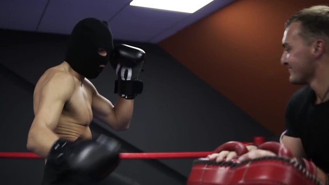 Aggressive boxer works with boxing paws with his coach in the ring. Practicing a series of leg punches. Shirtless athlete in black boxing gloves and thief mask. Slow motion
