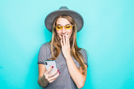 Portrait of outraged confused woman wearing fluppy hat and sunglasses holding and looking at mobile phone isolated over blue pastel background