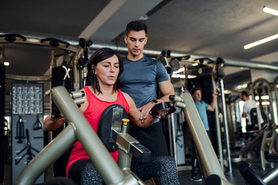 A female senior with a young trainer doing strength workout exercise in gym.