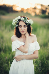 portrait of attractive brunette girl in white dress and with floral wreath in summer field at sunset