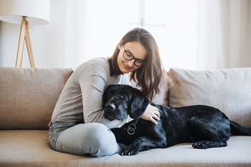 Teenage girl sitting on a sofa indoors, playing with a pet dog. - obrazy, fototapety, plakaty
