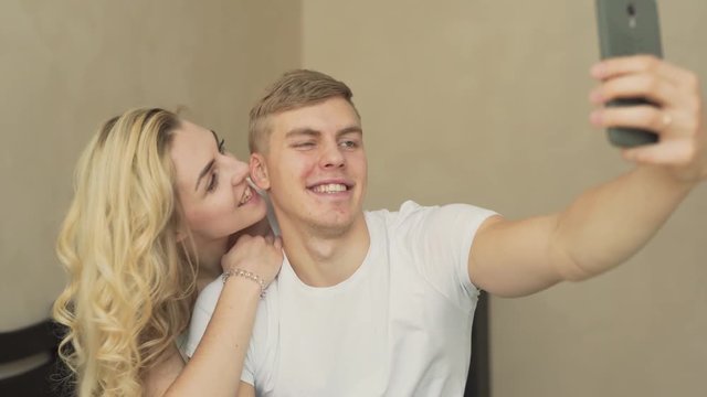 Cute couple is sitting on a bed in their apartment, hugging and making a selfie. Beautiful young woman is embracing her boyfriend at the back and smiling.