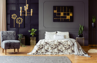Real photo of fresh plants and black and gold paintings hanging on walls with molding in dark bedroom interior with king-size bed and floral armchair - obrazy, fototapety, plakaty