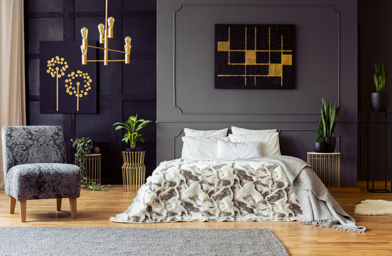 Real photo of fresh plants and black and gold paintings hanging on walls with molding in dark bedroom interior with king-size bed and floral armchair