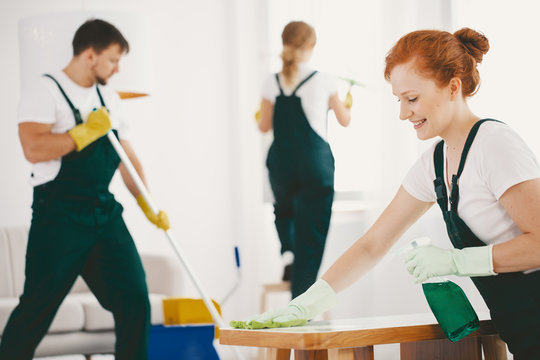 Smiling woman with gloves and spray wiping table while cleaning interior with crew