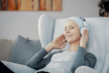 Peaceful woman with headphones and blue headscarf sitting in comfortable armchair resting at home...