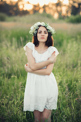 portrait of attractive brunette girl in white dress and with floral wreath in summer field at sunset